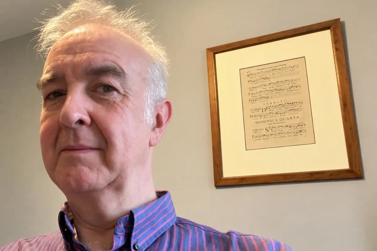 A man with short, spiky white hair and a striped shirt looks at the camera, with a framed piece of musical notation hanging on the wall behind him.