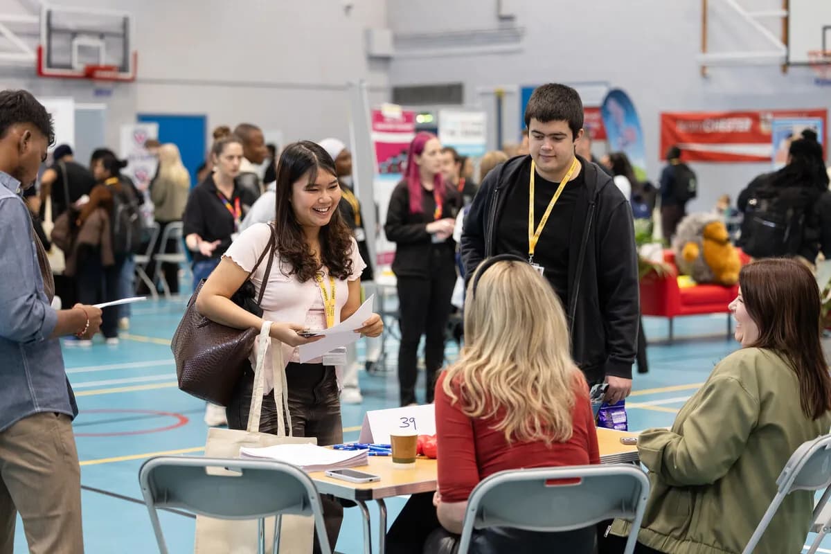 A young woman with long dark hair smiles while holding papers and a purse, interacting with people at a table in a busy hall.