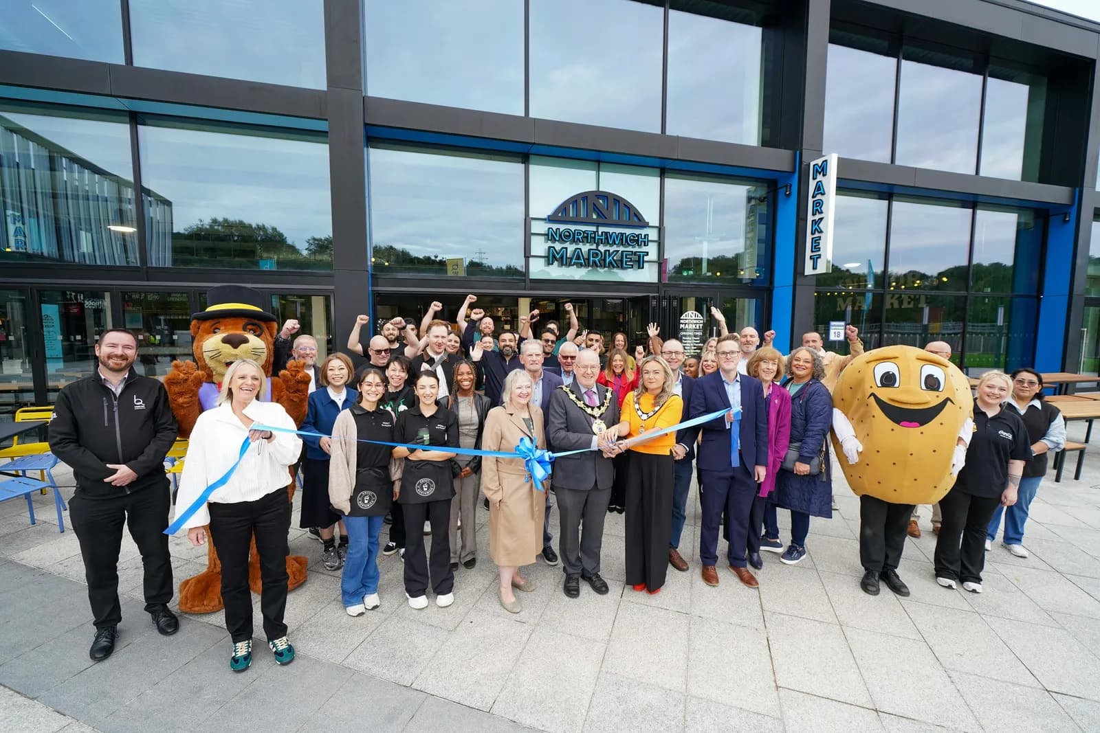 A diverse group of people, including mascots, gather in front of the Northwich Market building, holding a blue ribbon for a grand opening event.