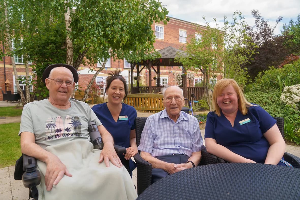 Two older men and two women in blue scrubs smile at the camera while sitting outside in a garden with trees, bushes, and a brick building in the background.