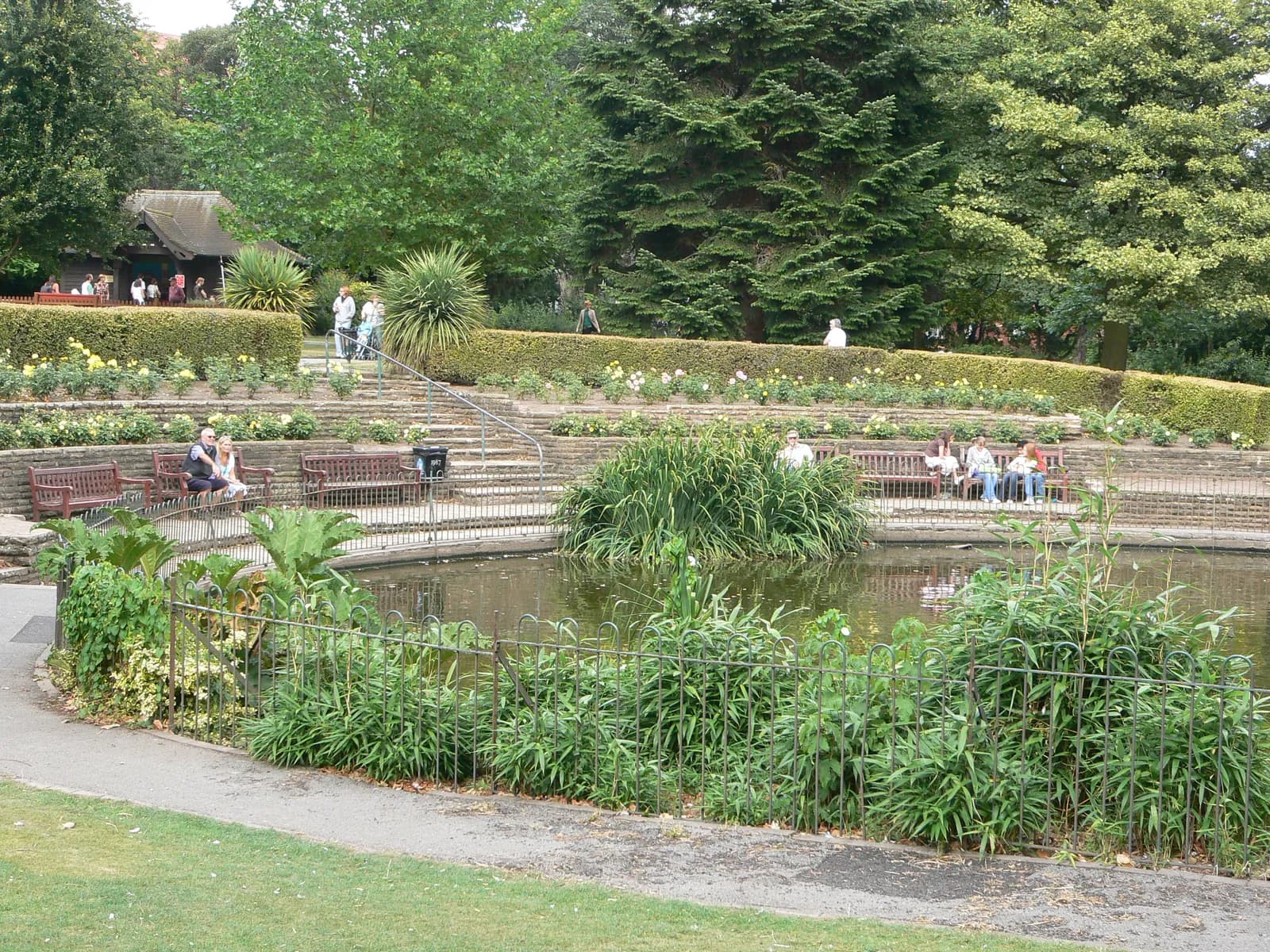 A terraced park with a small pond in the foreground, surrounded by lush green foliage and benches where people are seated.
