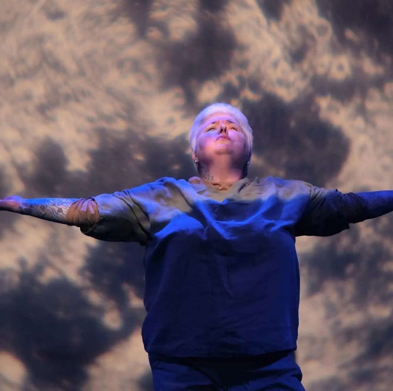 A person with short white hair and a blue and grey shirt stands with arms outstretched and head tilted back, against a cloudy backdrop.